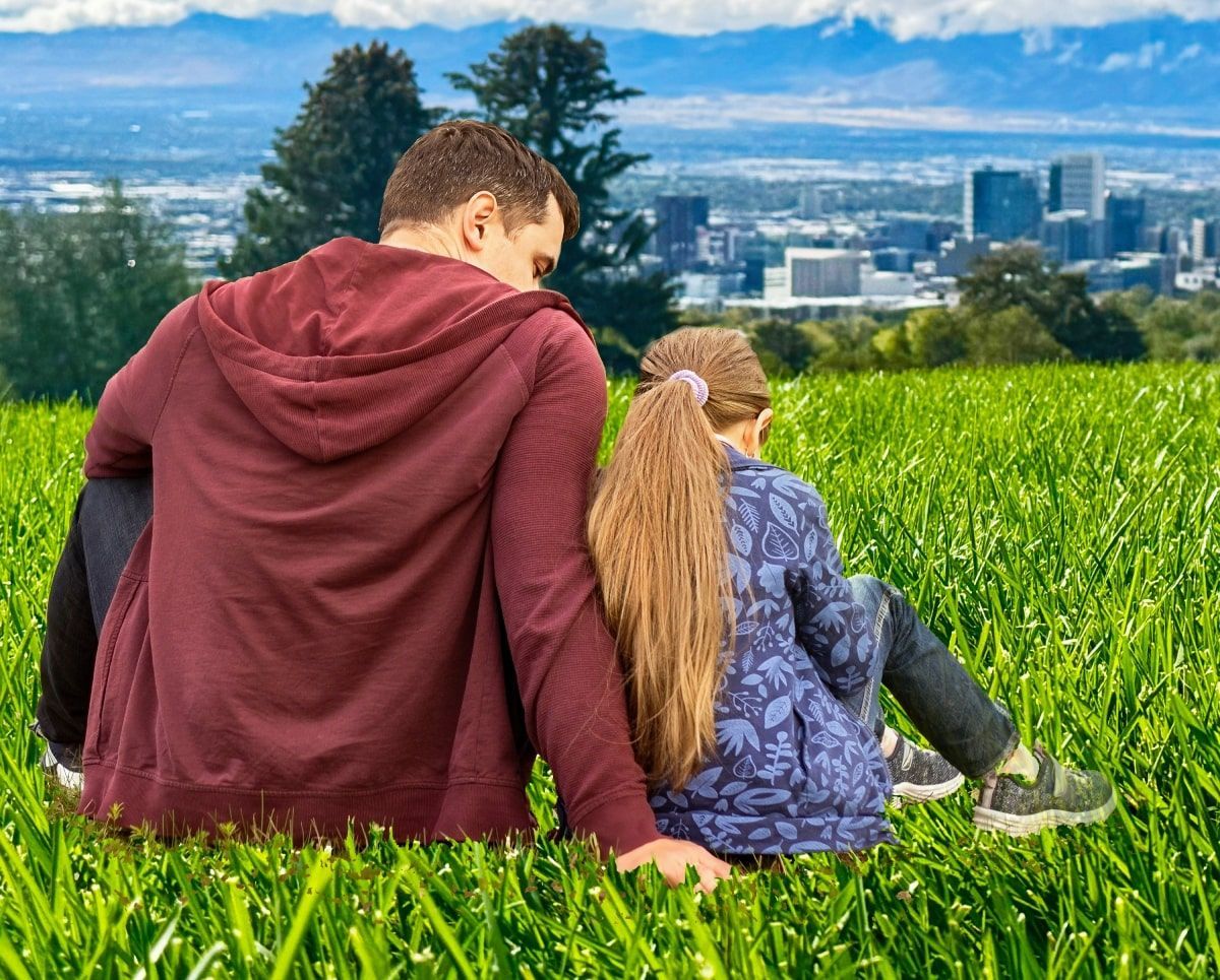 Man and child sitting on a grassy hill, overlooking a city.