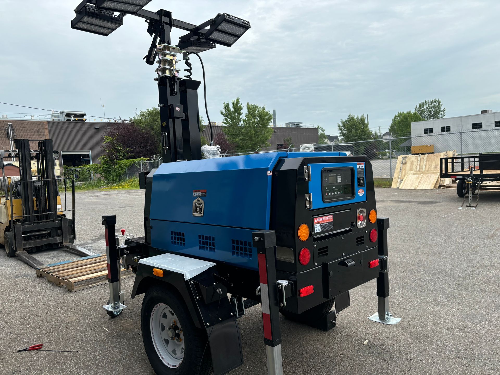 Des projecteurs de chantier illuminent une rue la nuit. Un gros générateur bleu se trouve au premier plan, avec des maisons à l'arrière-plan.