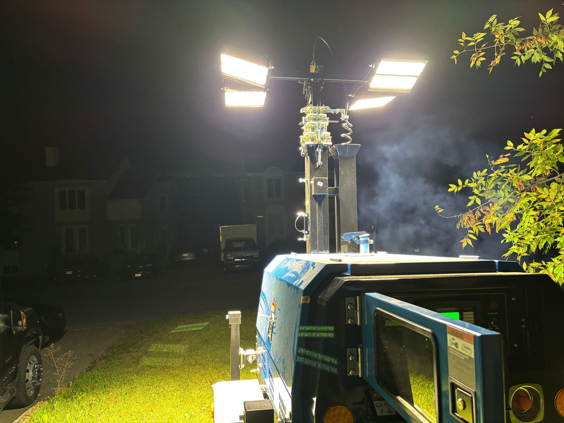 Des projecteurs de chantier illuminent une rue la nuit. Un gros générateur bleu se trouve au premier plan, avec des maisons à l'arrière-plan.