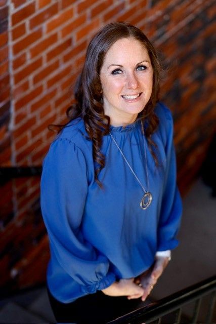 A woman in a blue shirt is standing in front of a brick wall.
