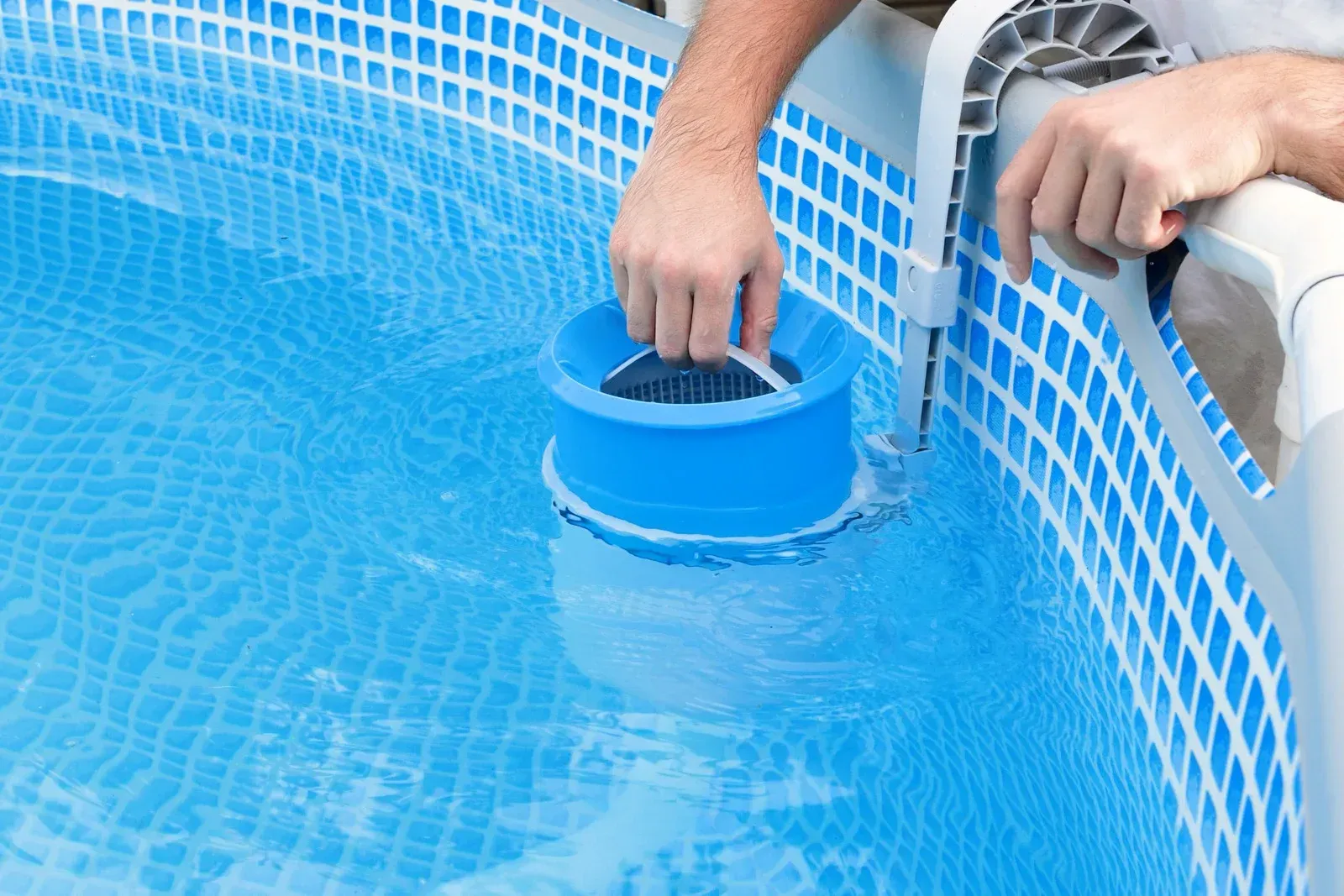Person cleaning an above-ground pool skimmer, blue pool, and white and blue tiled walls.