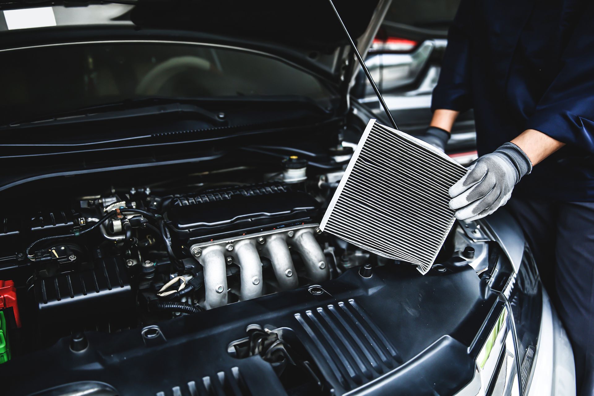 Gloved hands holding a rectangular air filter above an open car engine during routine maintenance. Gloved hands holding a rectangular air filter above an open car engine during routine maintenance.