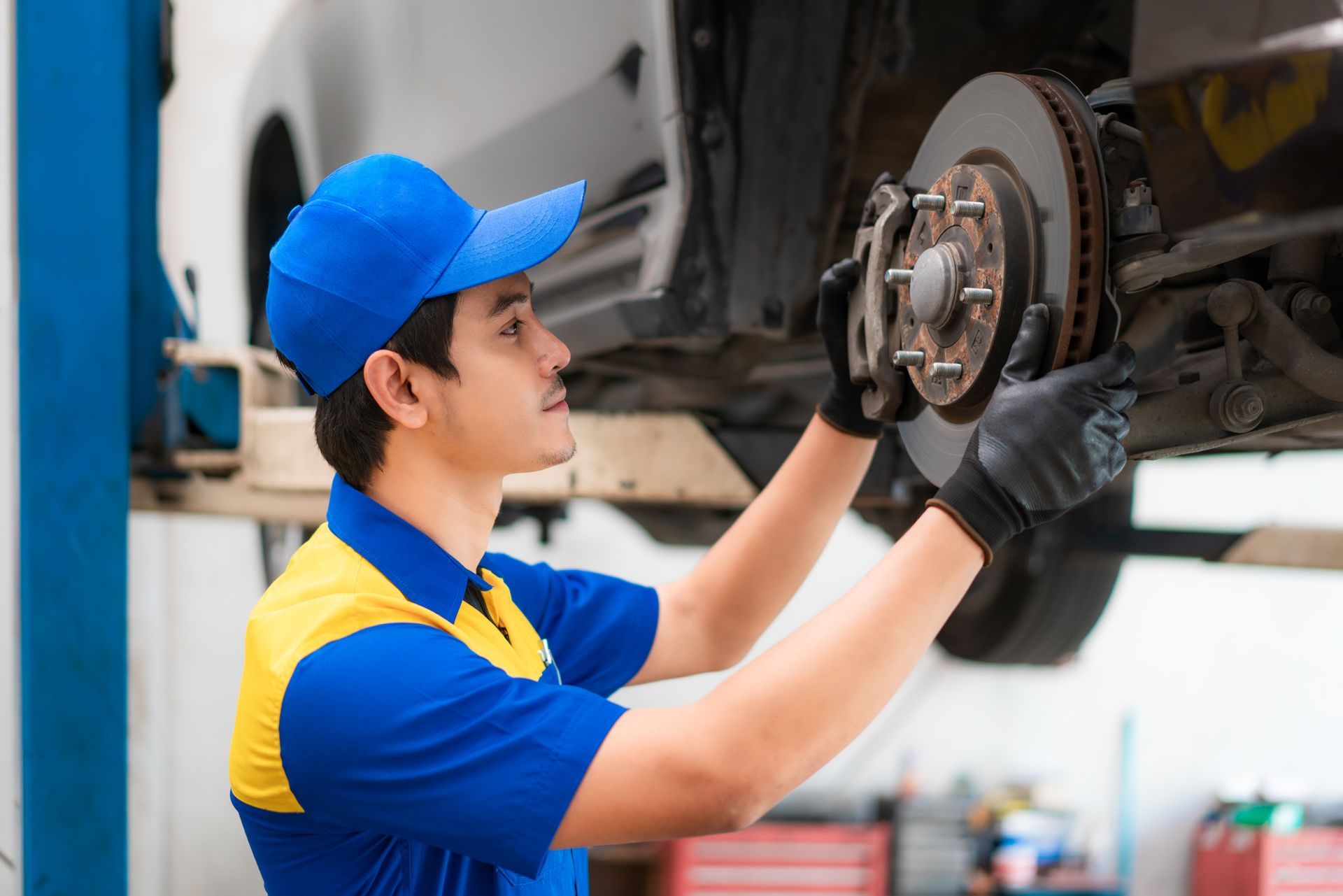Mechanic inspecting brake parts on lifted car in auto repair shop wearing blue uniform.
