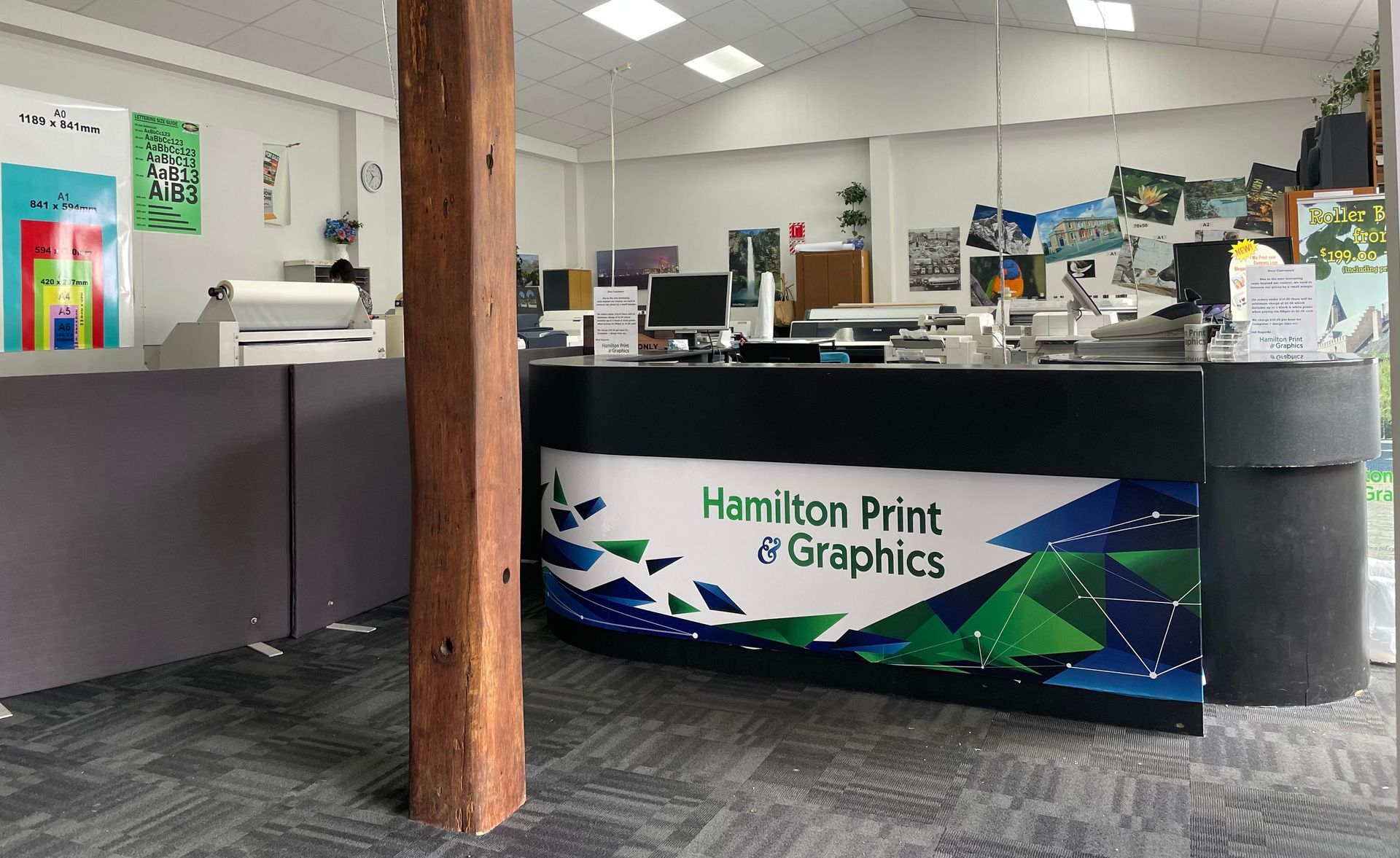 Interior of Hamilton Print & Graphics office, featuring a reception desk with logo, posters, and equipment.