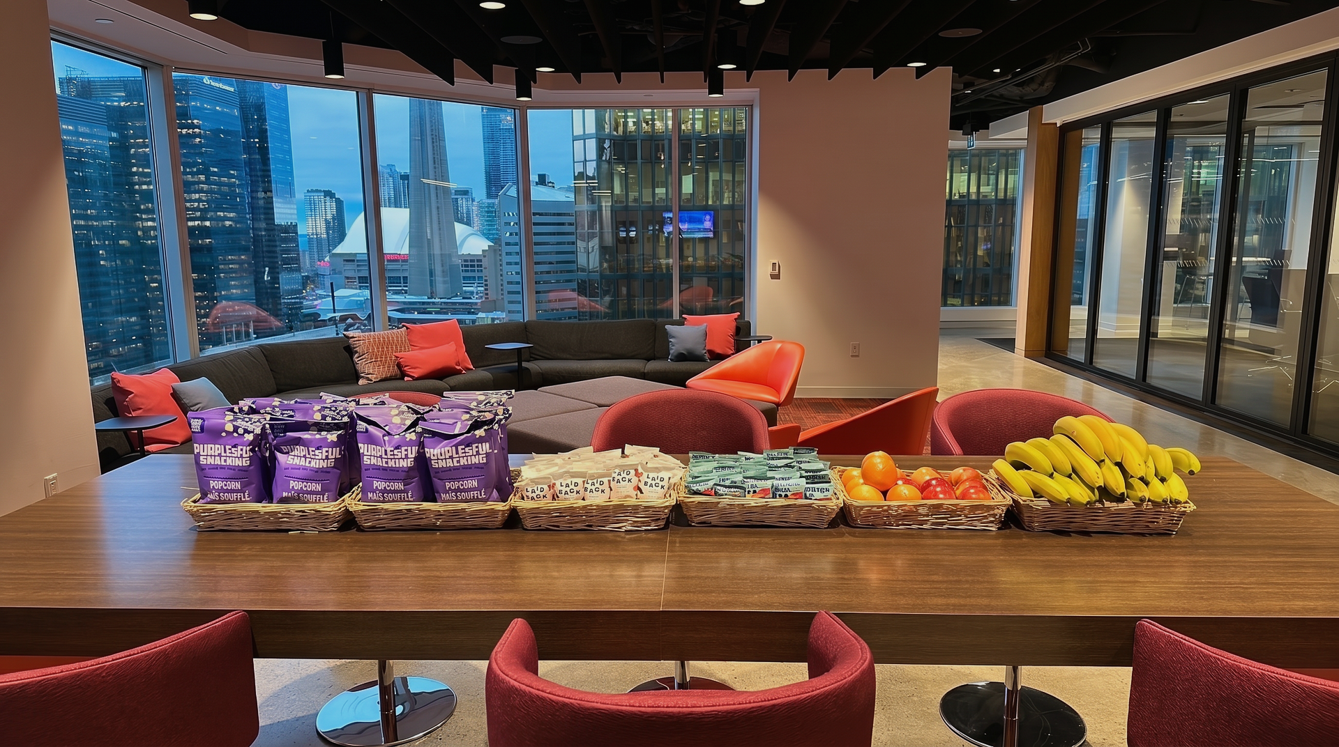 Modern lounge with a long snack display of pastries, fruit, and wrapped treats by large windows.