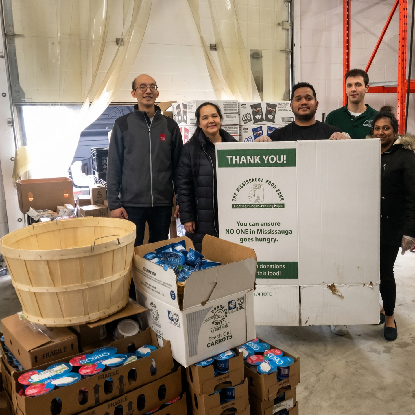 Volunteers stand beside donated food boxes and a large “Thank You!” sign in a warehouse.