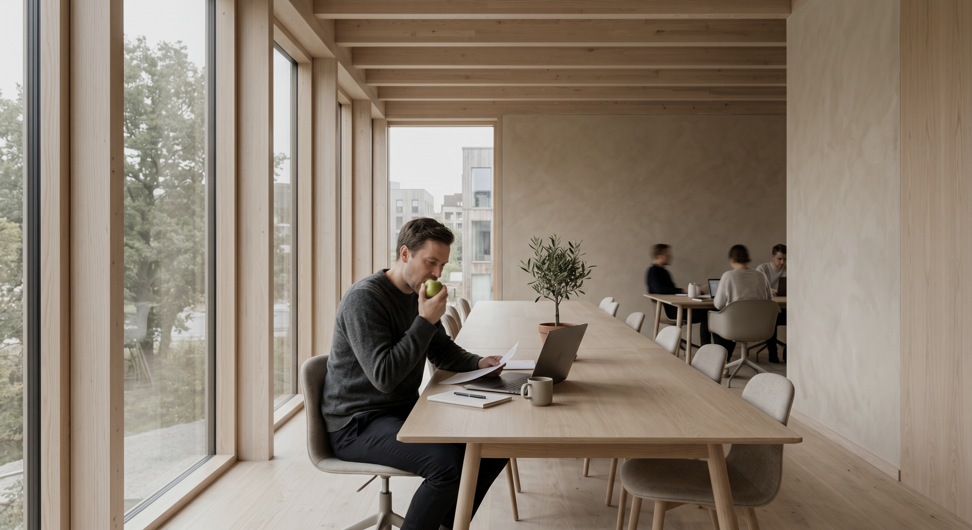 Modern office lounge with people working at a long wooden table by large windows