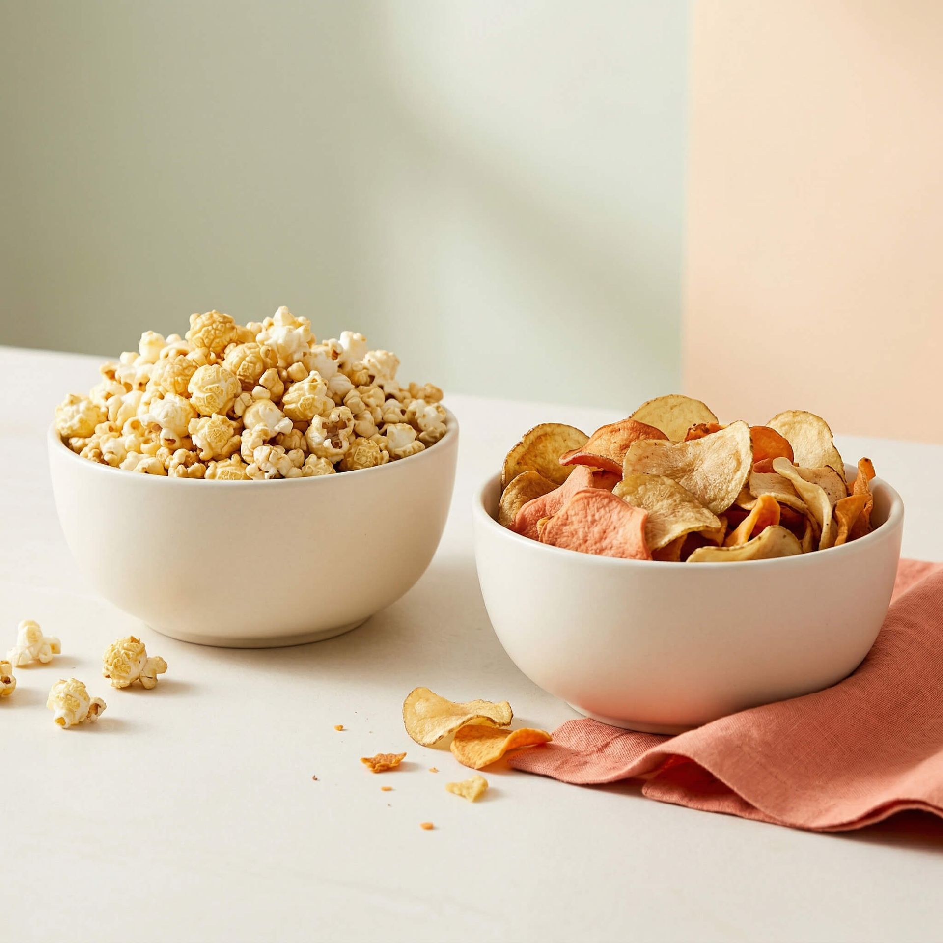 Two white bowls of popcorn and potato chips on a light surface with a pink cloth