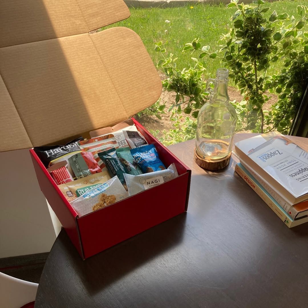 Open red gift box with assorted snacks on a table beside a glass and stacked books by a sunny window