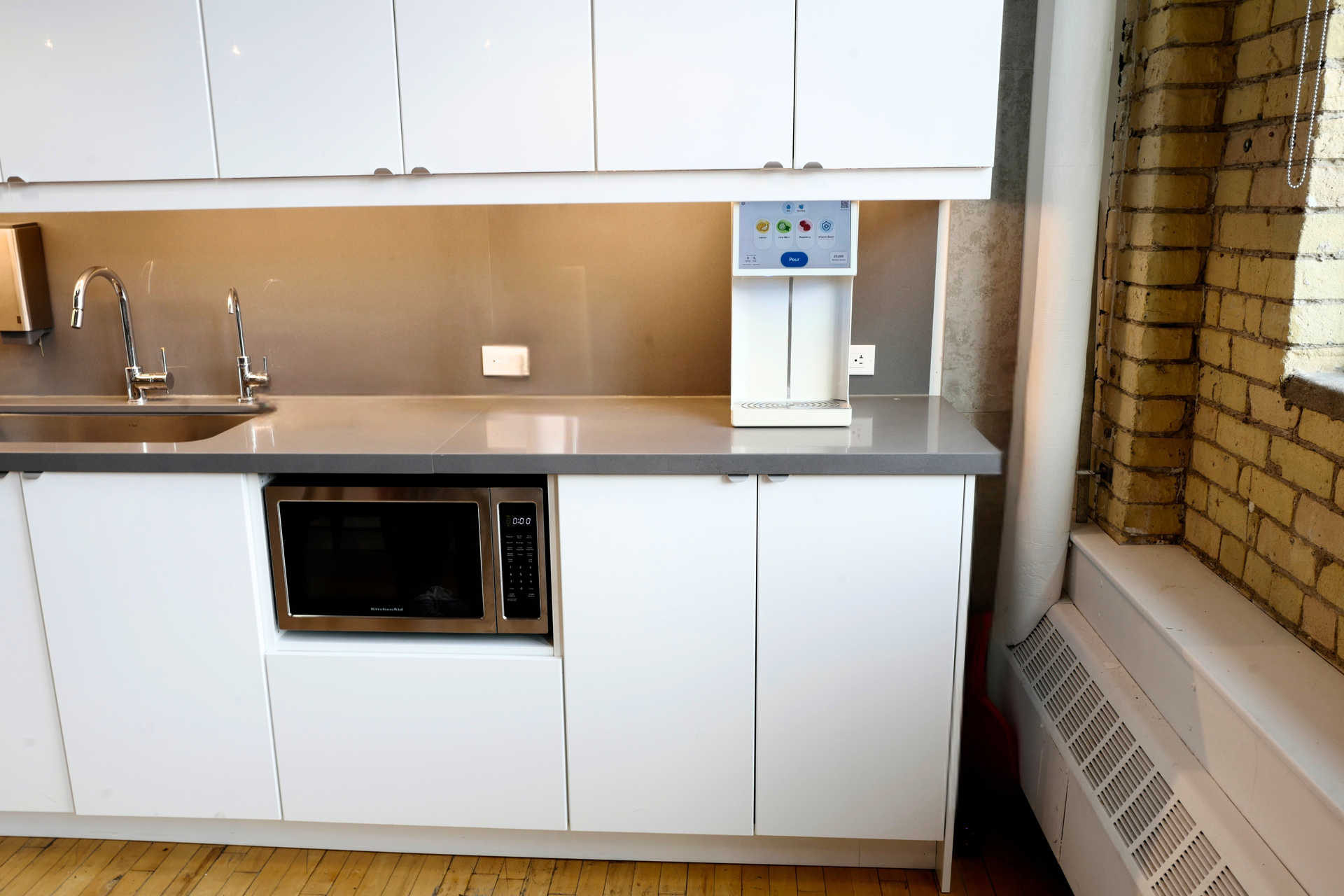 Modern kitchen with white cabinets, stainless countertop, sink, microwave, and water dispenser by brick wall