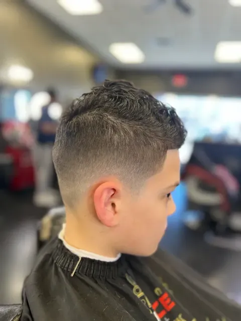 Boy with a short fade haircut sitting in a barbershop chair, wearing a black cape.