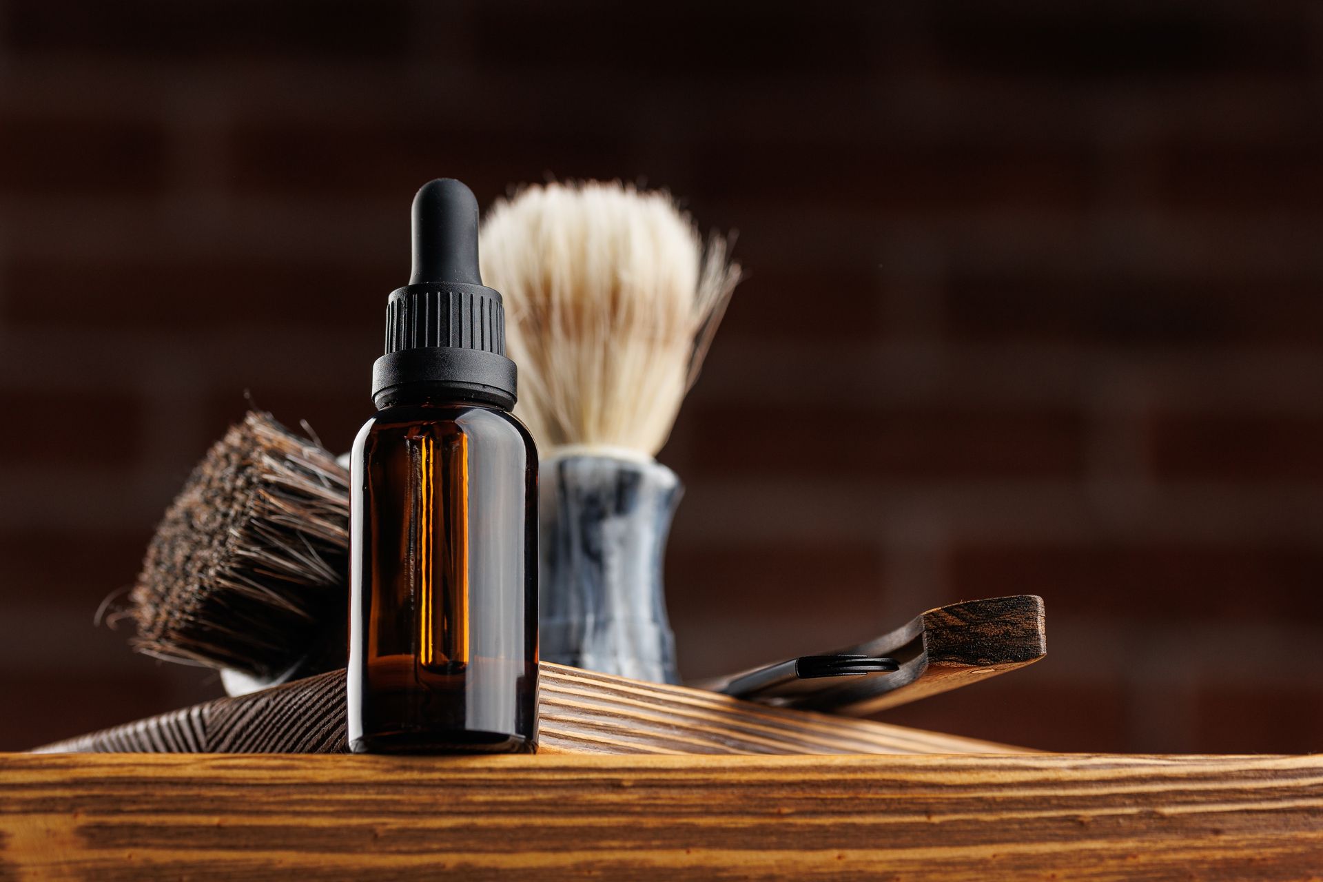 Barber tools on a wooden table: amber bottle, brush, and comb against a brick background