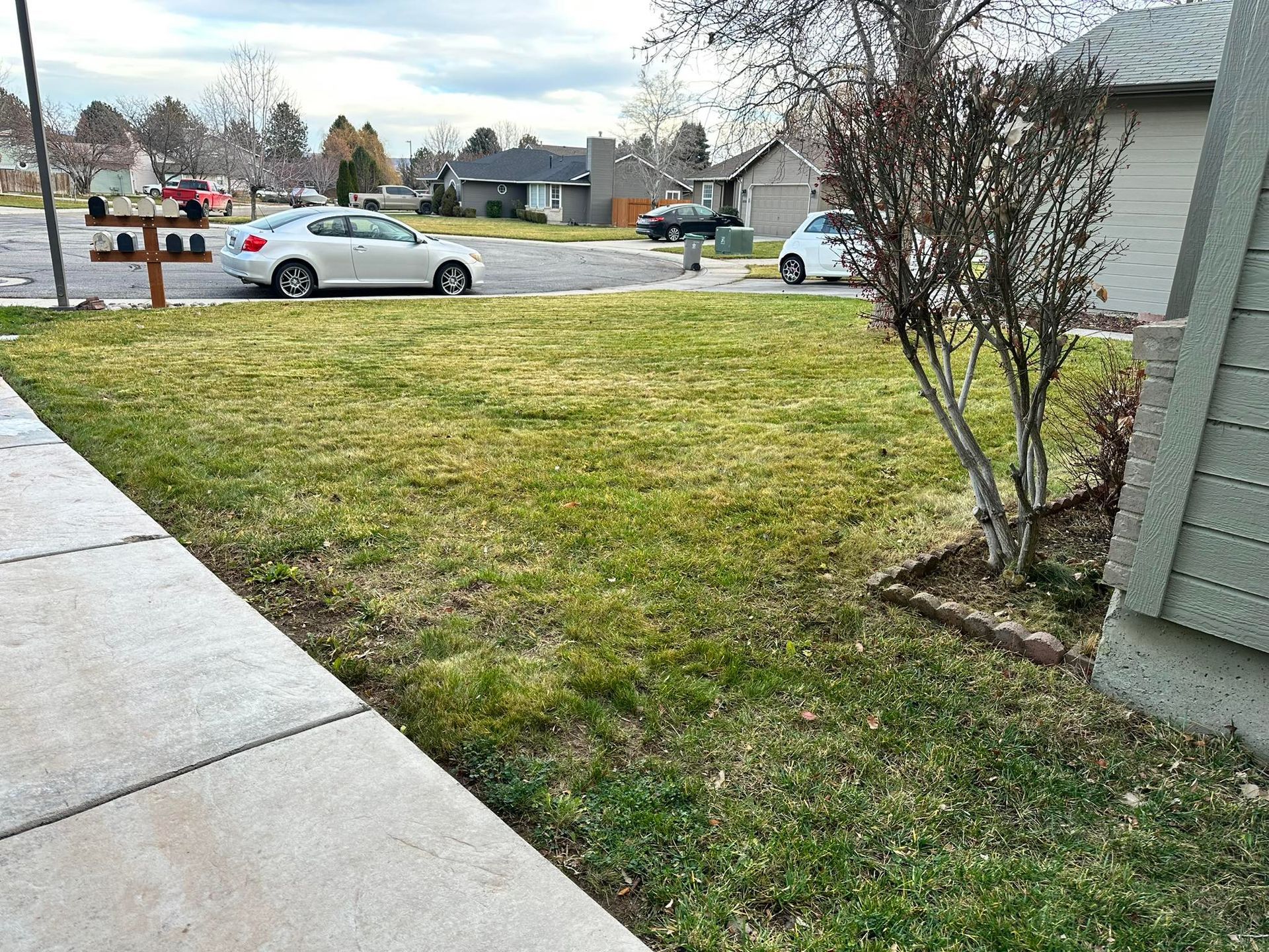 A view from a sidewalk of a green lawn in a suburban neighborhood with a white car parked on the street.