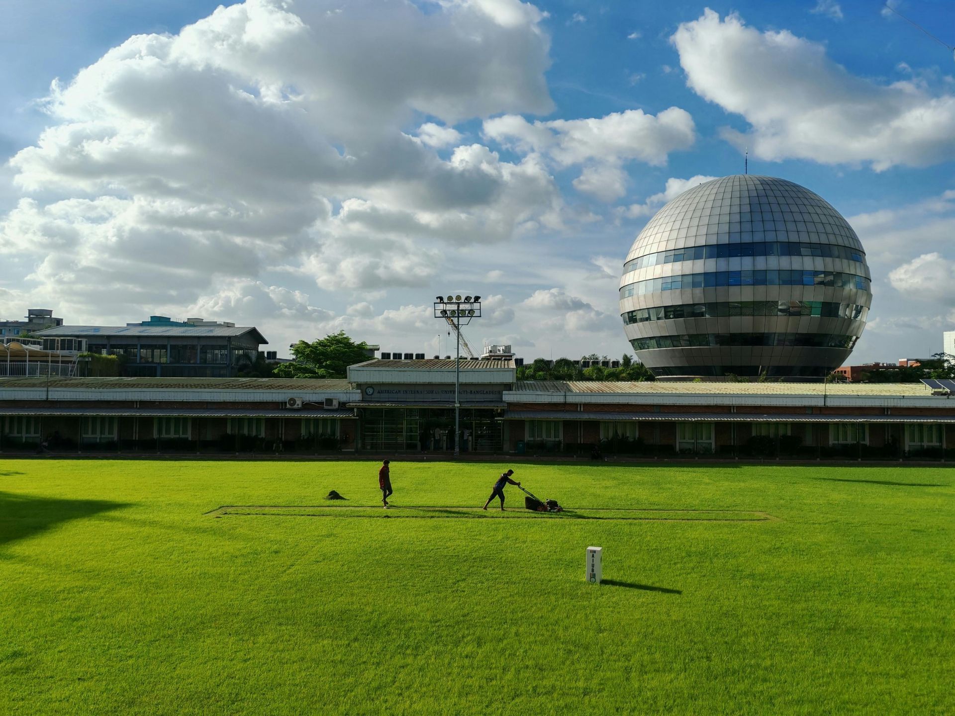 Two people maintain a vibrant green lawn in front of a long building and a prominent spherical, metallic structure.