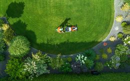 An overhead view of a person pushing a lawnmower across a lush green lawn surrounded by garden beds.