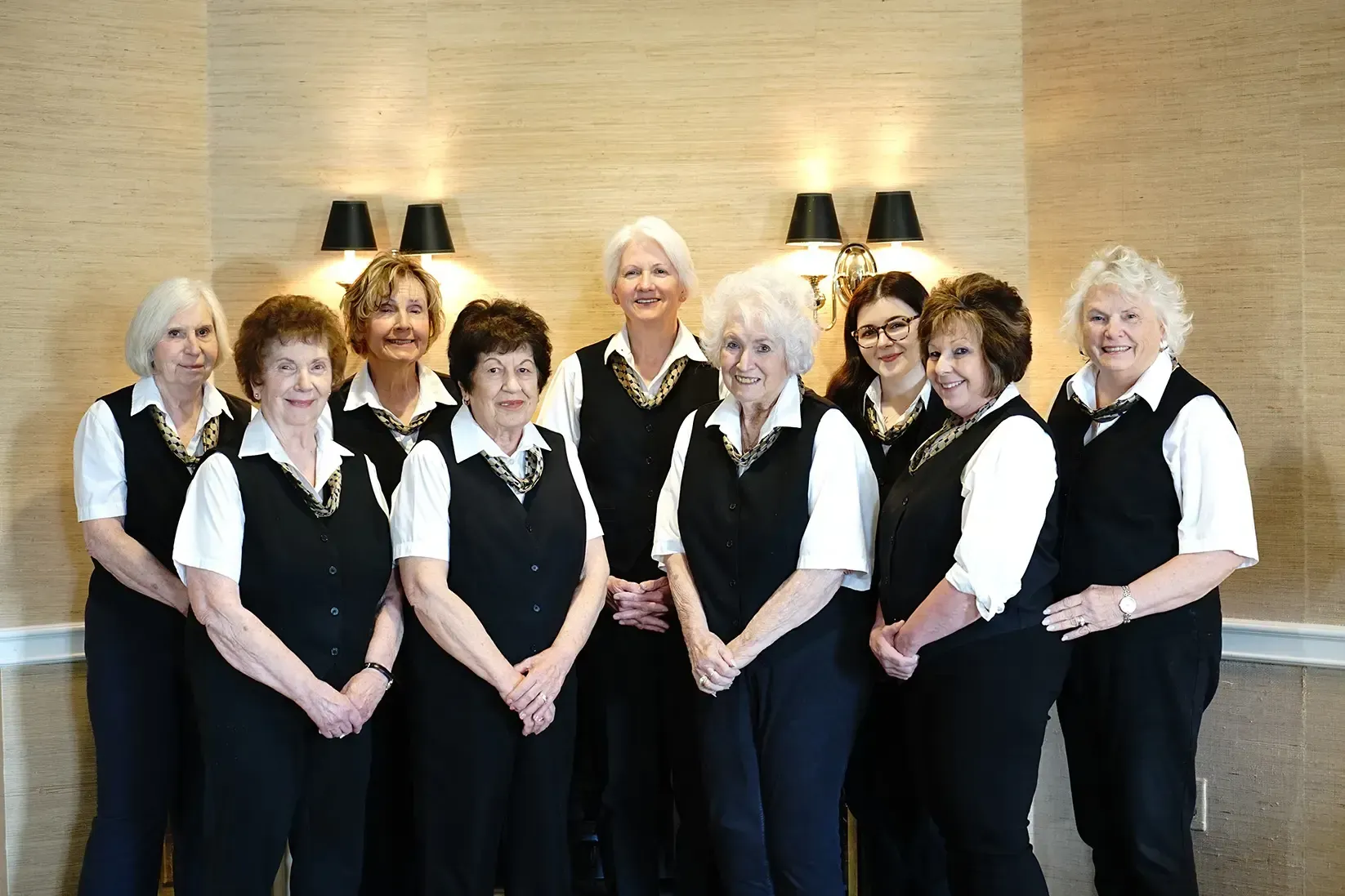 Eight women in black-and-white uniforms standing together indoors, smiling in front of a stone wall.
