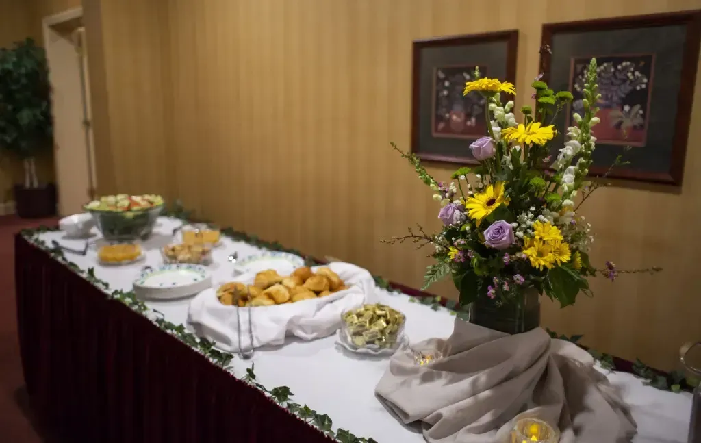 Buffet table with white tablecloth, platters of food, and a large yellow and purple flower arrangement
