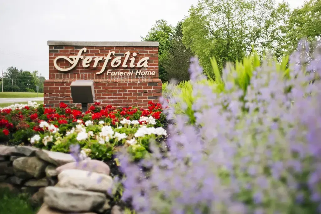 Ferfolia Funeral Home sign amid red flowers, rocks, and soft purple blossoms in a landscaped garden