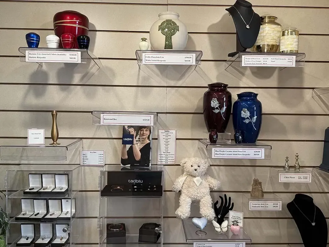 Shelf display of assorted cremation urns and keepsake items, including teddy bears and jewelry, in a shop.