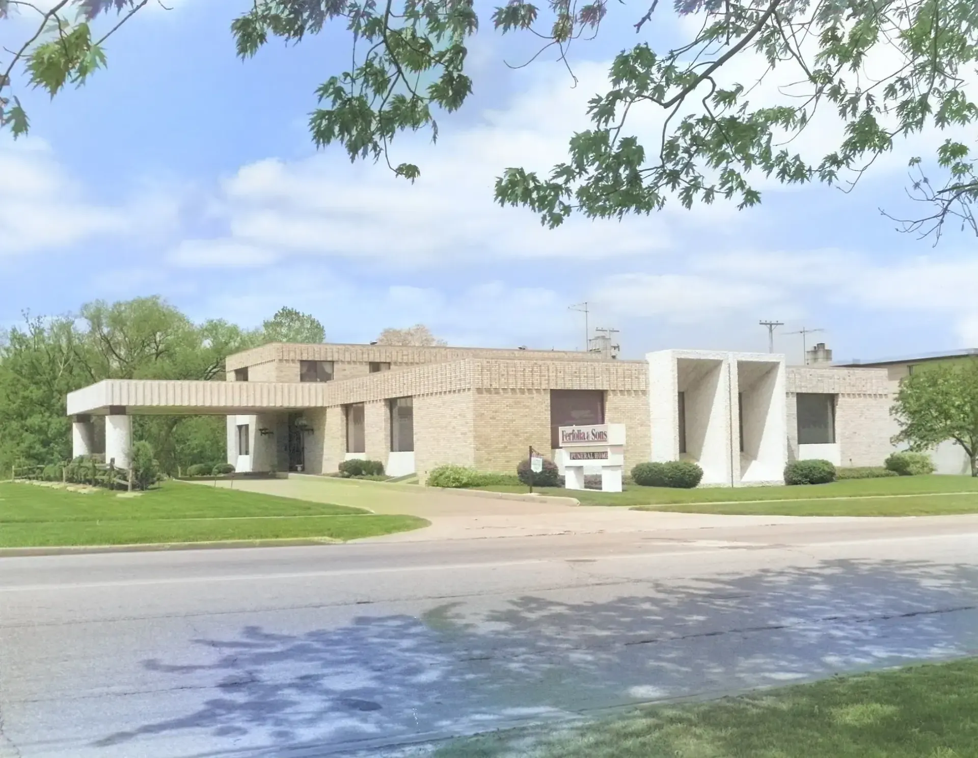 Low stone building with white columns beside a grassy lawn and road under a leafy sky.