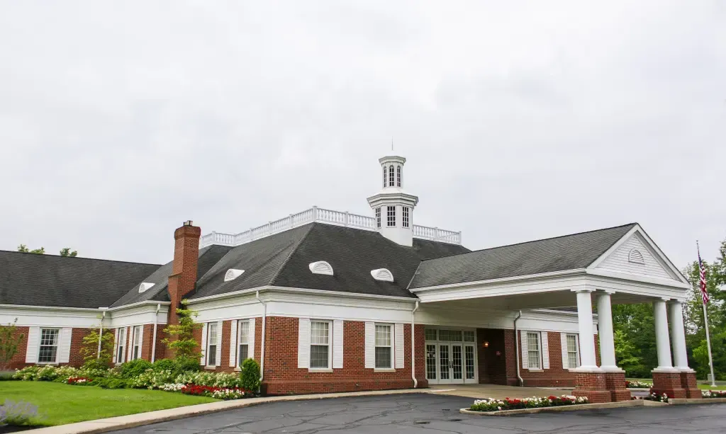 Brick clubhouse with white columns and cupola, landscaped entrance under an overcast sky