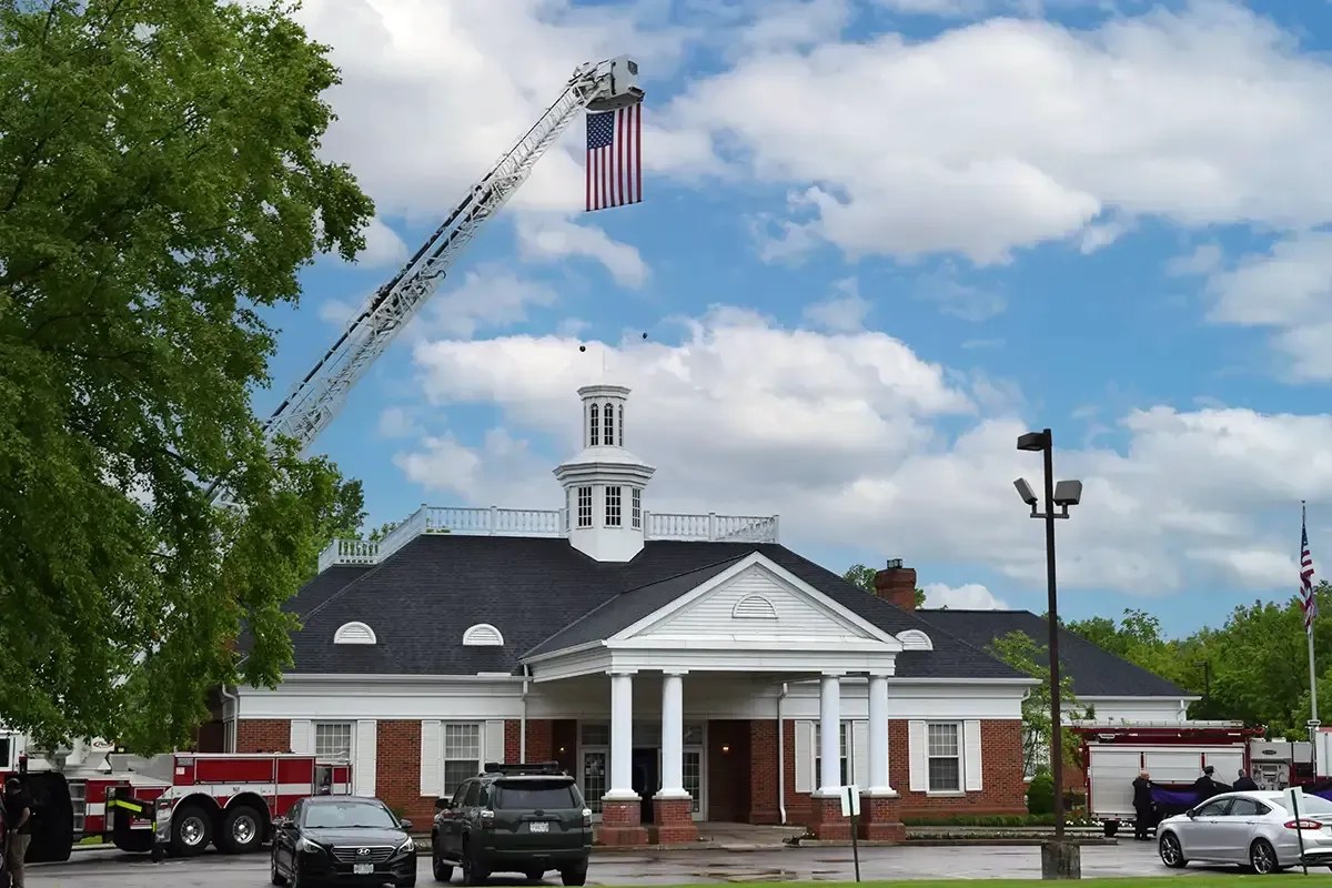 Brick civic building with white columns and a U.S. flag, with a fire truck ladder raised beside it in the rain