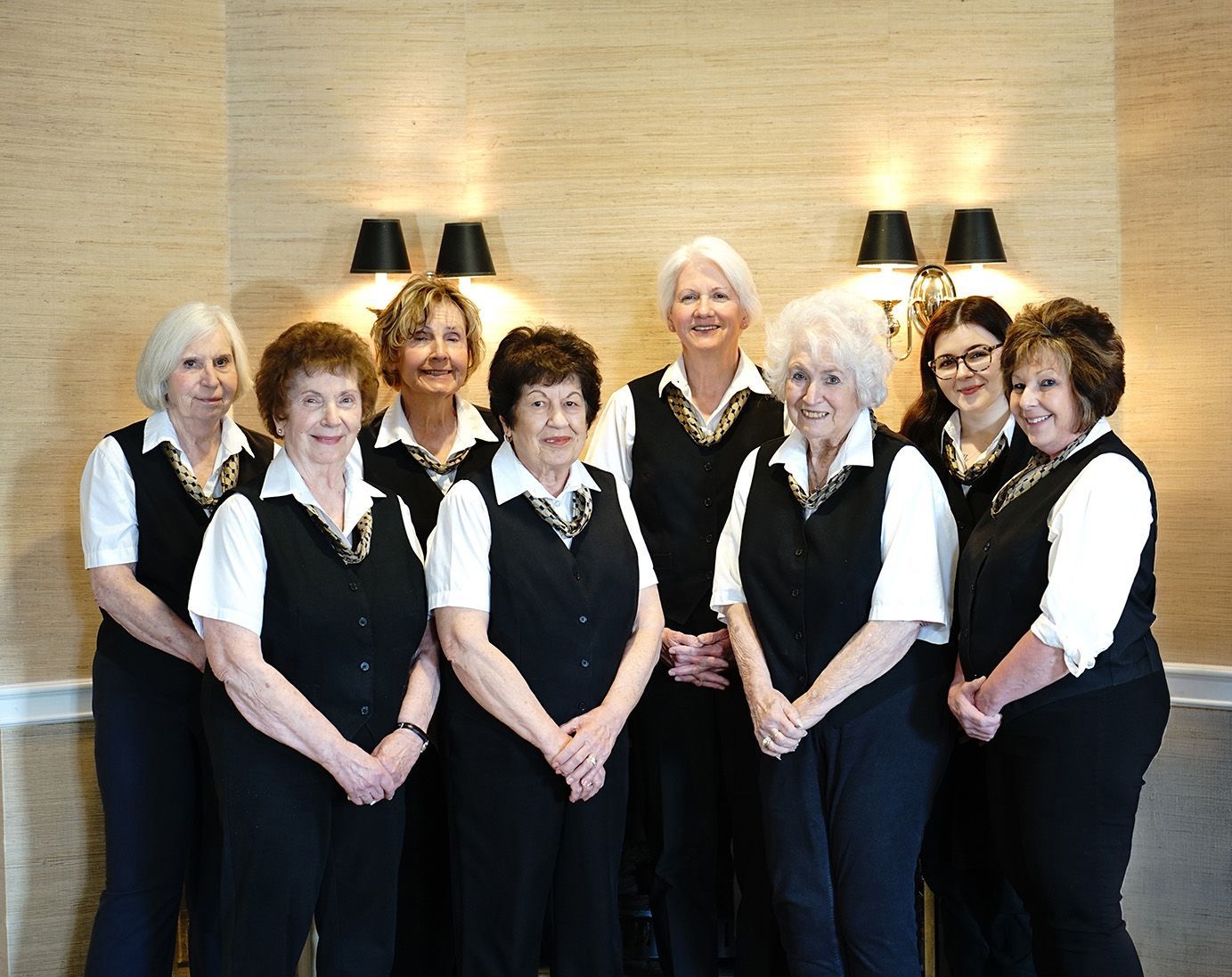 Eight women in black-and-white uniforms standing together indoors, smiling in front of a stone wall.