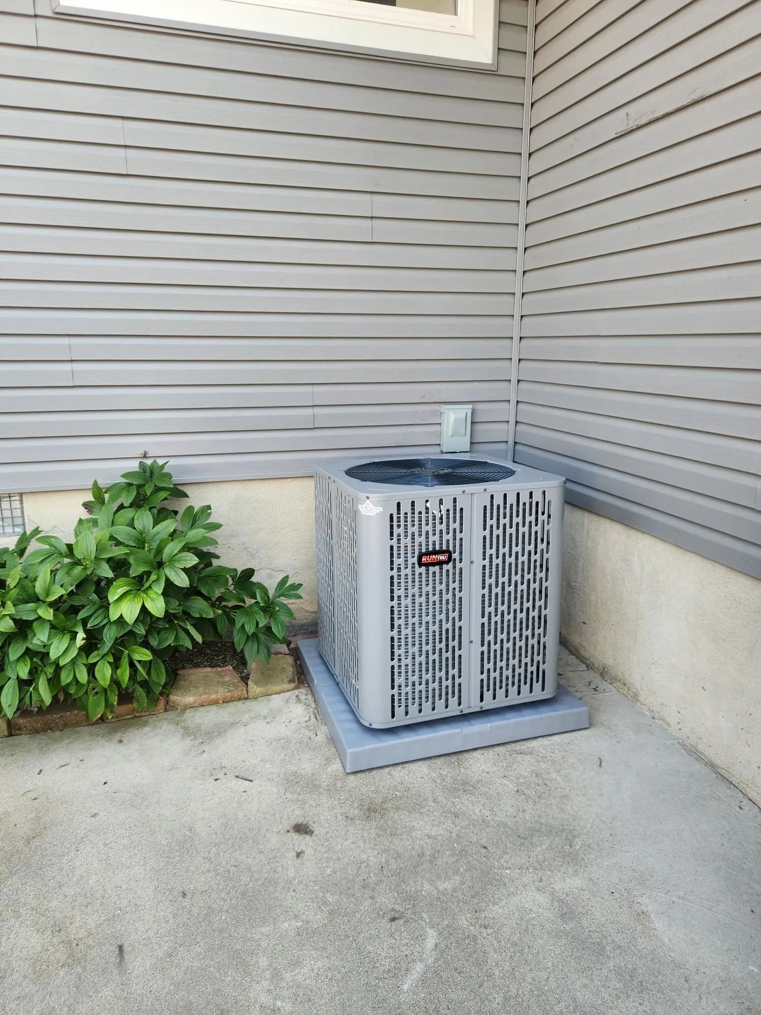 Outdoor air conditioner unit beside house siding and a bush on a concrete patio