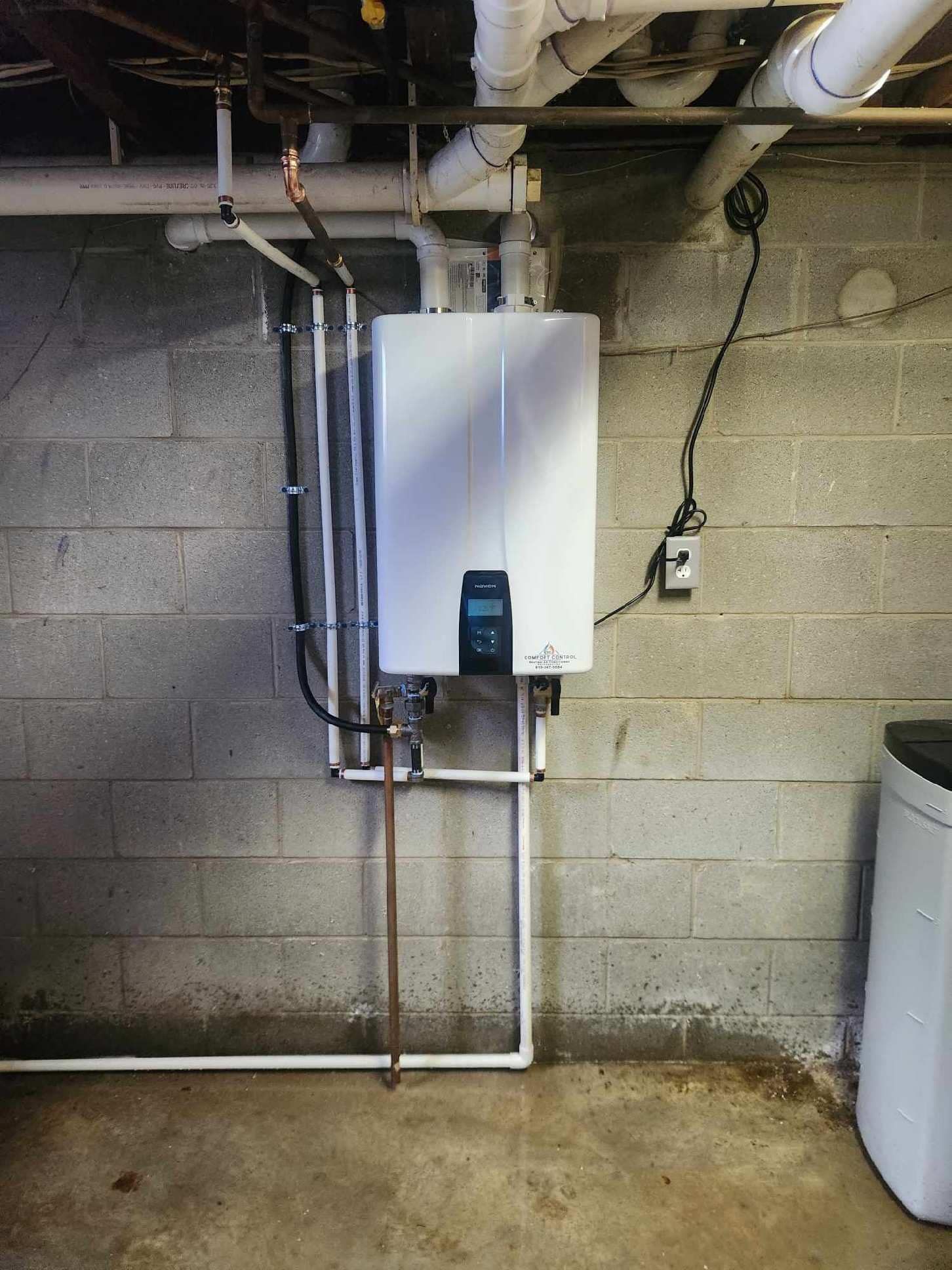 Tankless water heater mounted on a concrete block wall in a basement, surrounded by pipes.