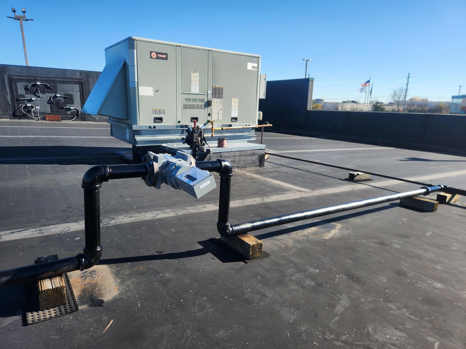 HVAC unit on rooftop with black piping, valve, and metal conduit. Sunny day.
