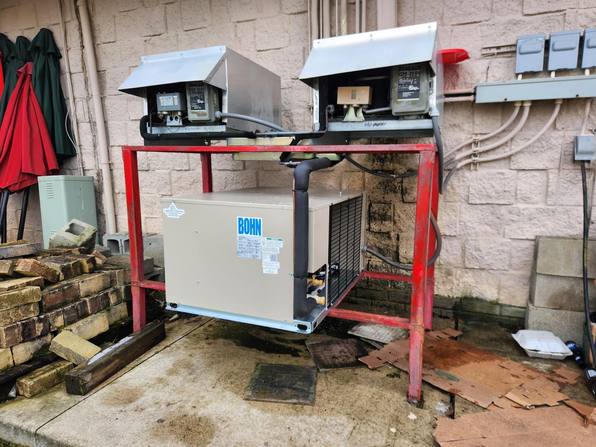 Outdoor HVAC system on red metal frame, with two metal-boxed meters above and brick wall backdrop.
