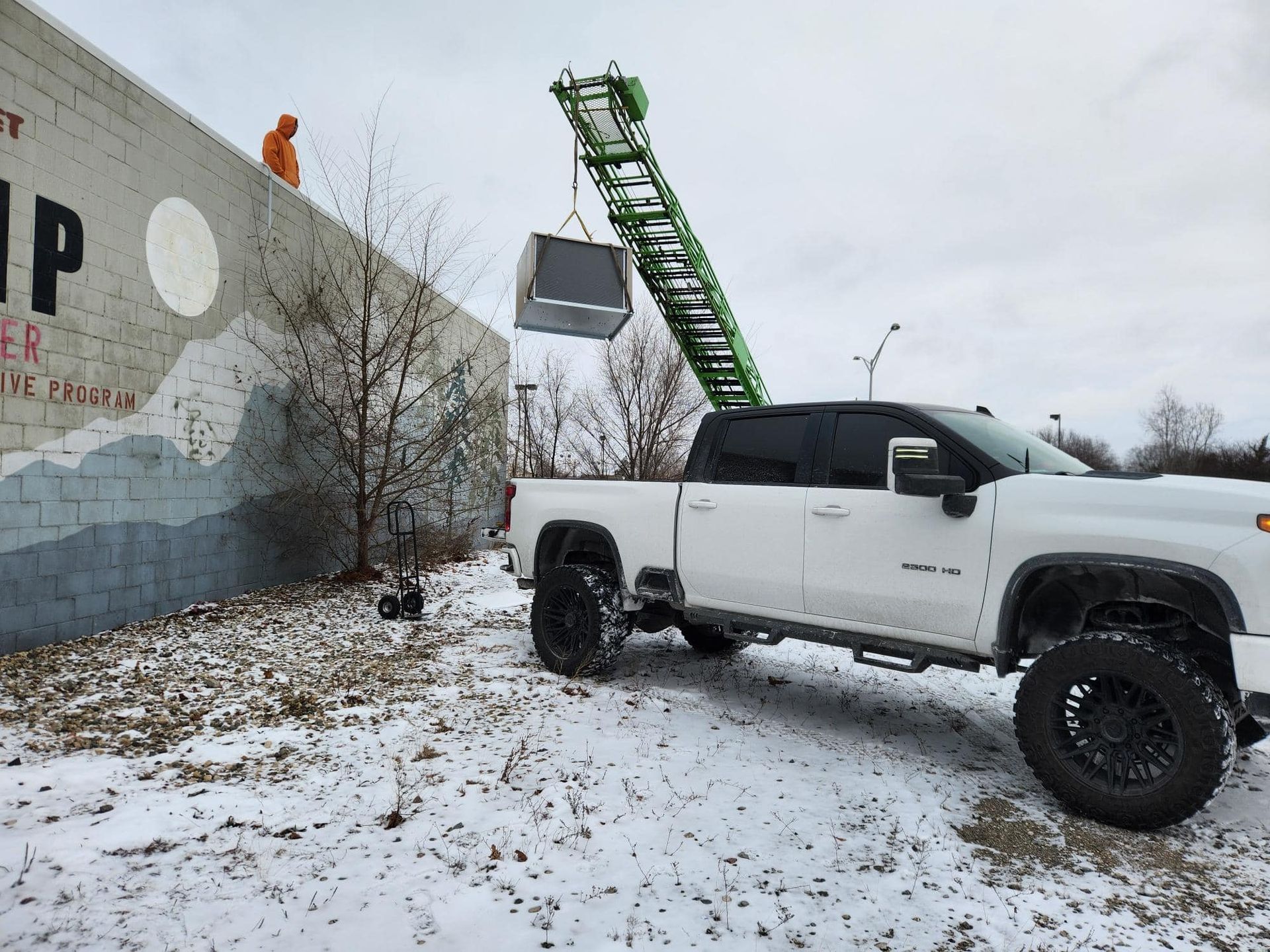 White truck using a lift to move a gray box over a wall on a snowy day. A person watches from the wall.