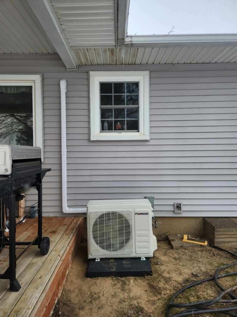 An outdoor HVAC unit sits near a house's exterior wall with a window and a covered porch.