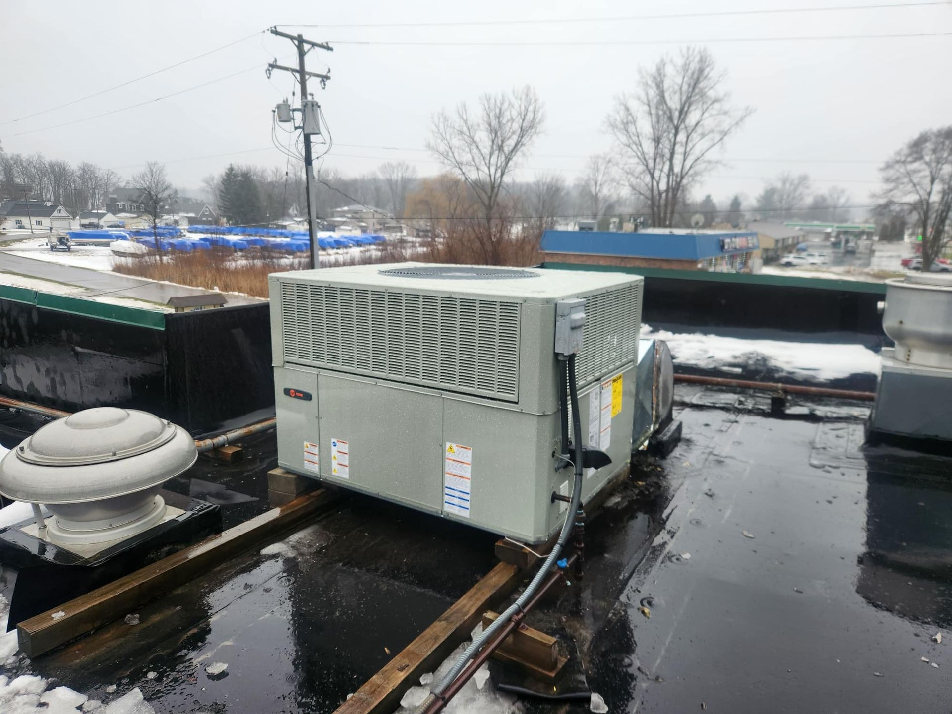 HVAC unit on a flat commercial roof with snow. Gray unit, overcast day, wooden supports.