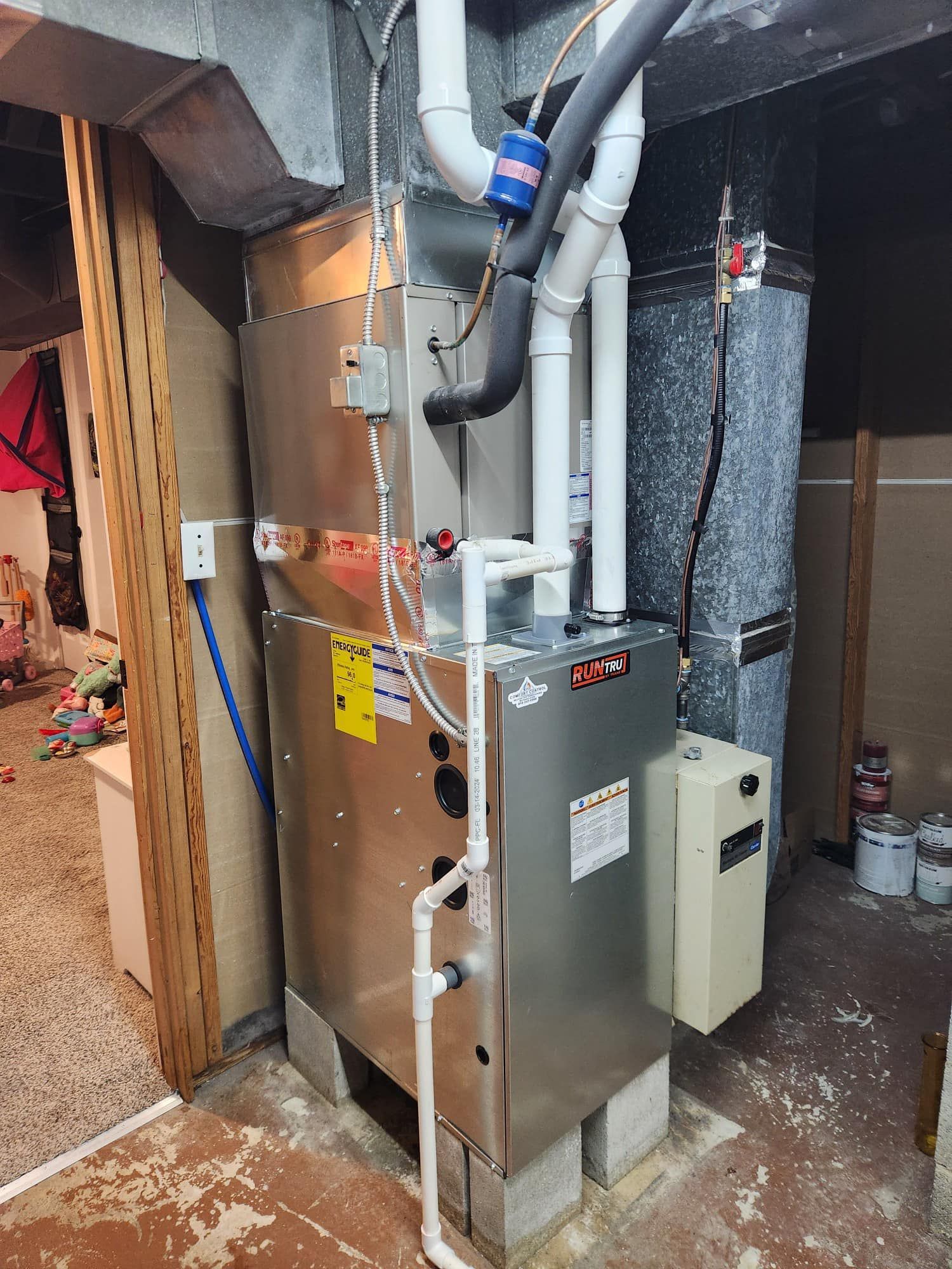 A furnace and related components in a basement. Silver metal unit with white pipes and ductwork.