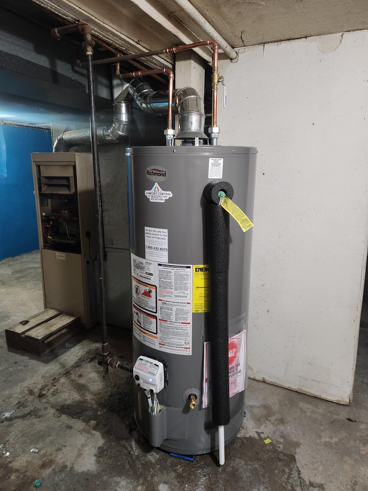 A tall, gray water heater in a basement next to a furnace. Copper pipes run overhead.
