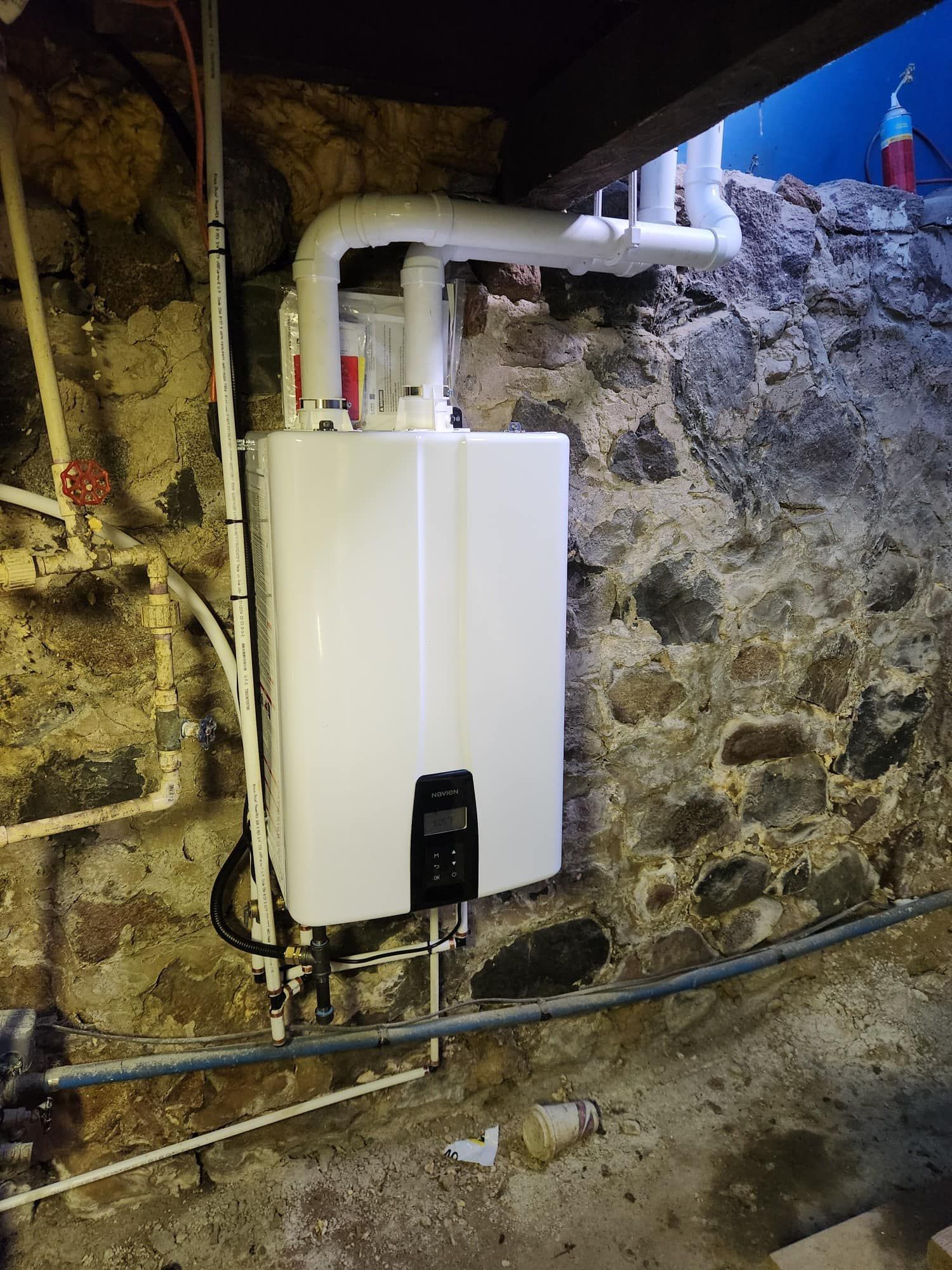 Tankless water heater installed against a stone wall in a basement. White unit with connected pipes.