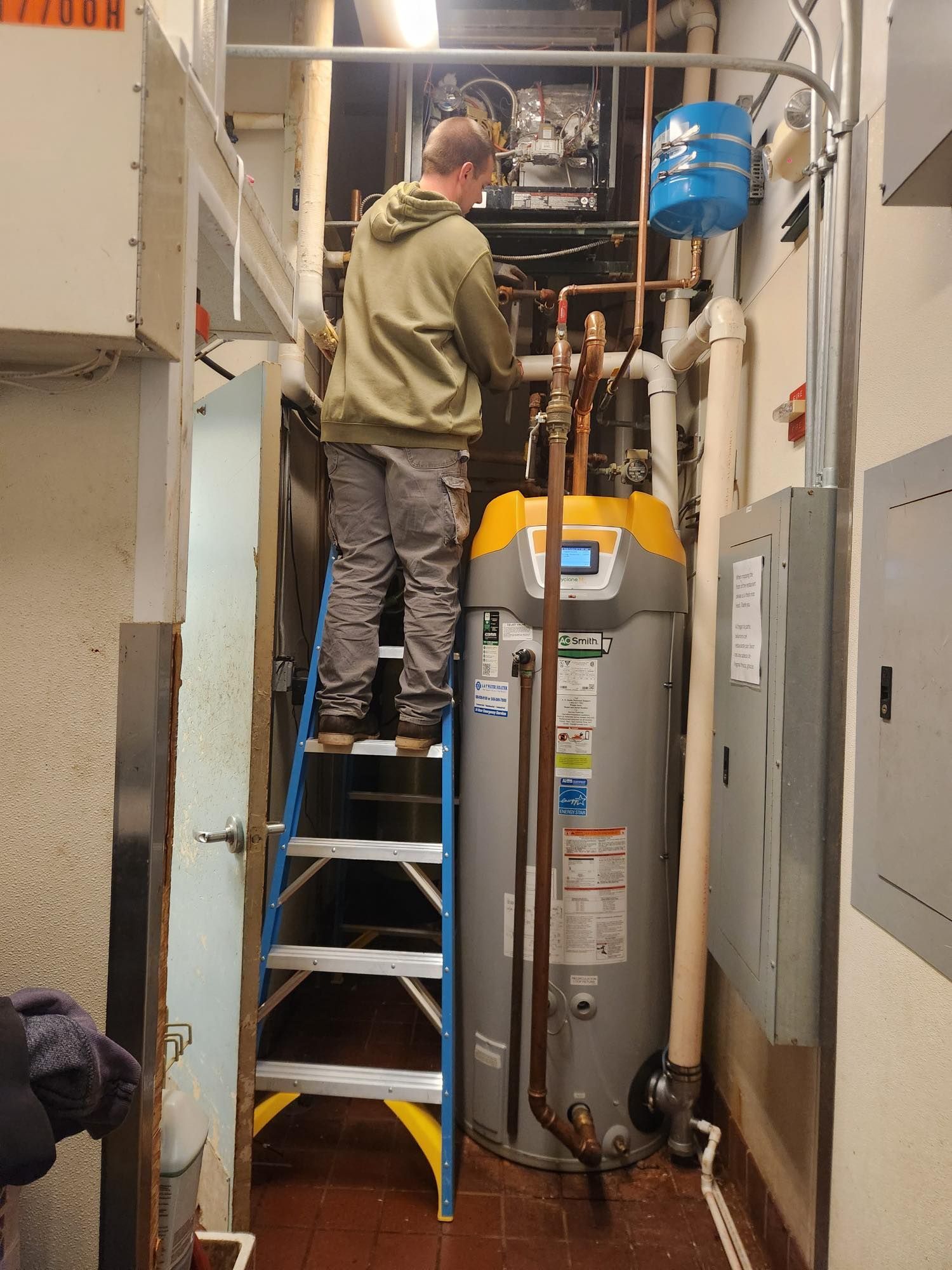 Person on a ladder working on pipes near a water heater and water pressure tank in a utility room.