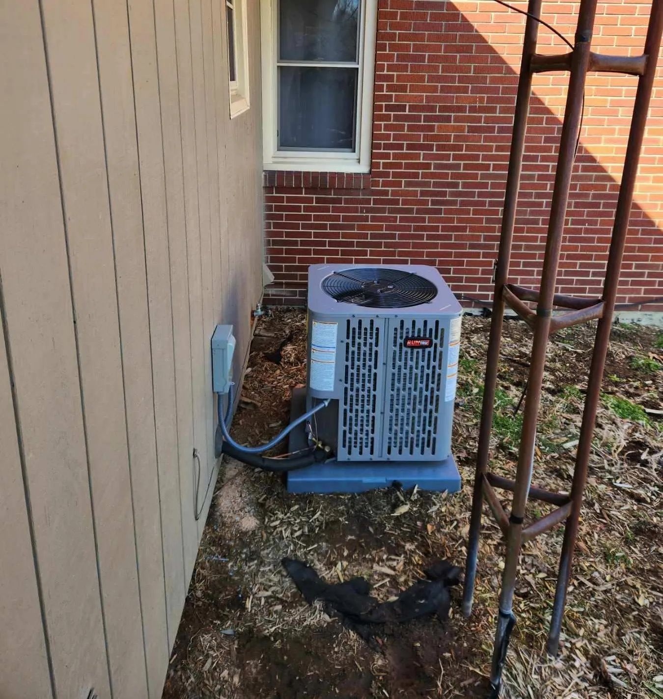 Outdoor air conditioner unit beside a beige wall and brick house, with black metal trellis in front.