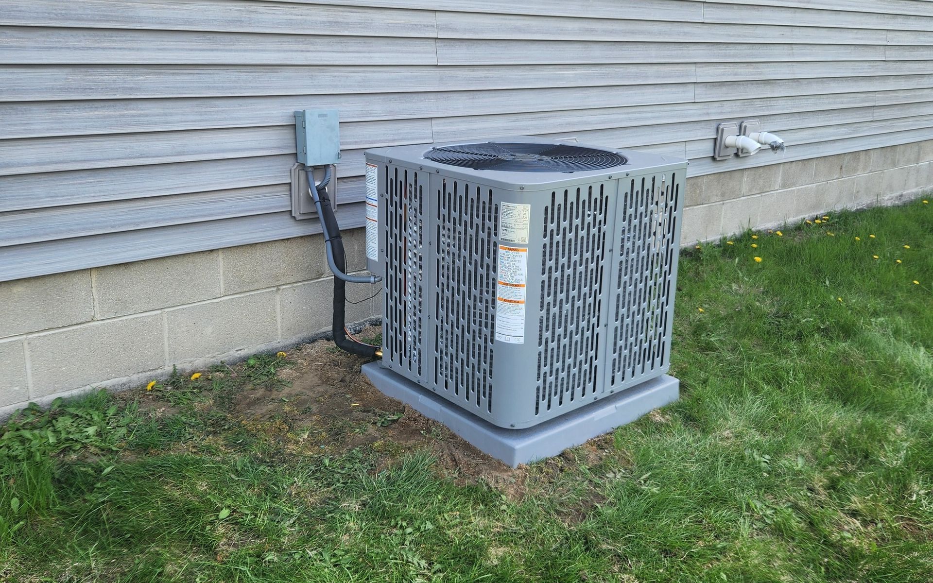 AC unit outside a home, gray with a metal grate, sitting on a concrete pad with electrical components nearby.