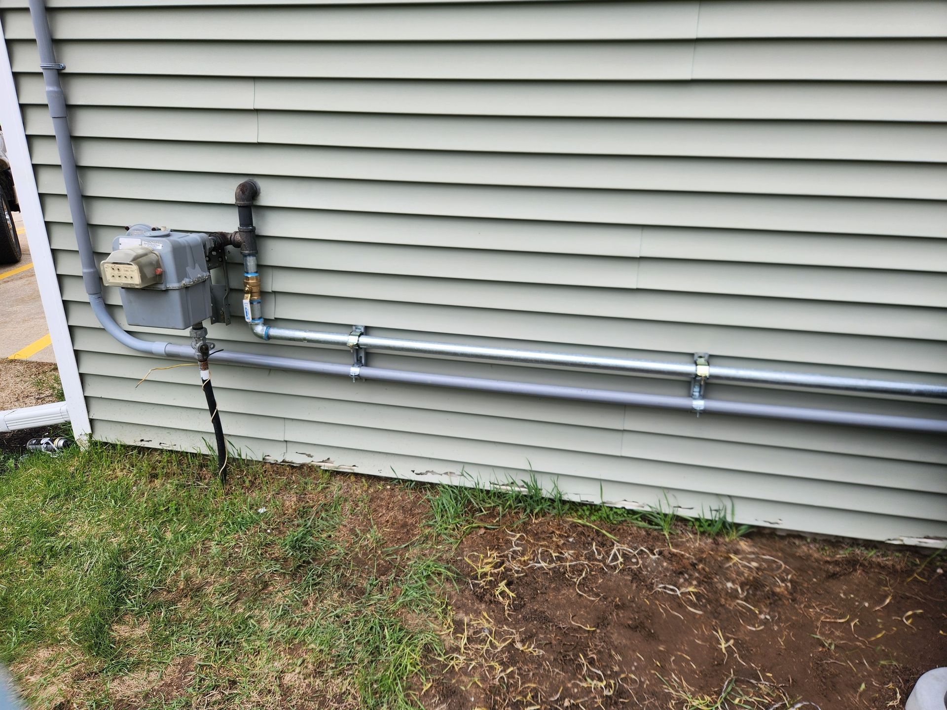 Gas meter and piping on a green-sided building. Silver pipes are secured with metal clamps on the wall. 