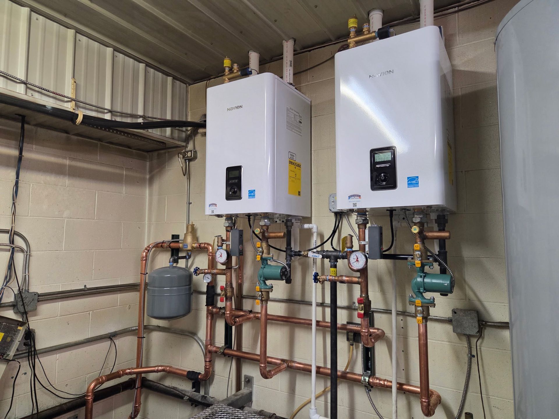 Two white wall-mounted water heaters with copper pipes, gauges, and a gray expansion tank, in an equipment room.