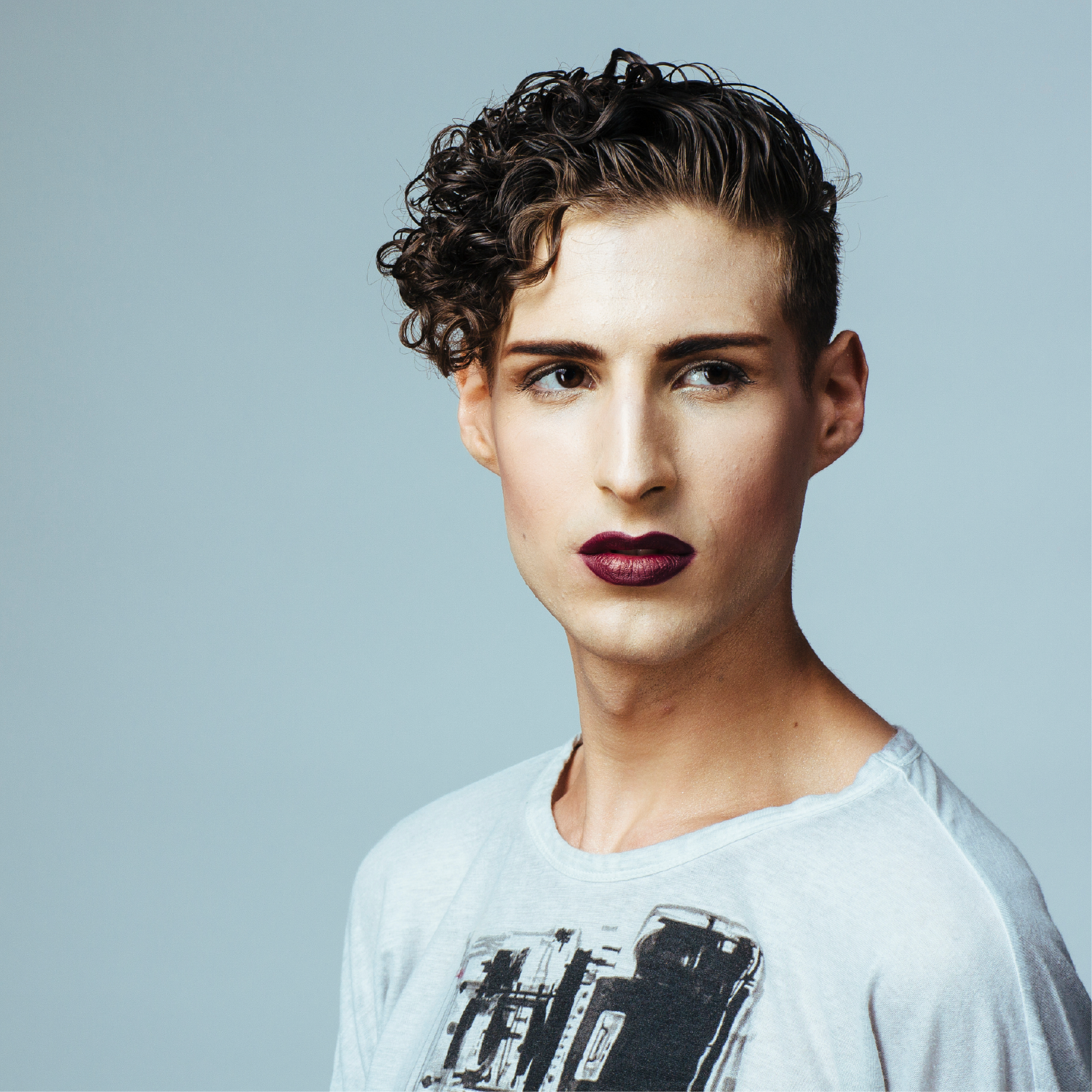 A young man with curly hair and purple lipstick is wearing a gray shirt
