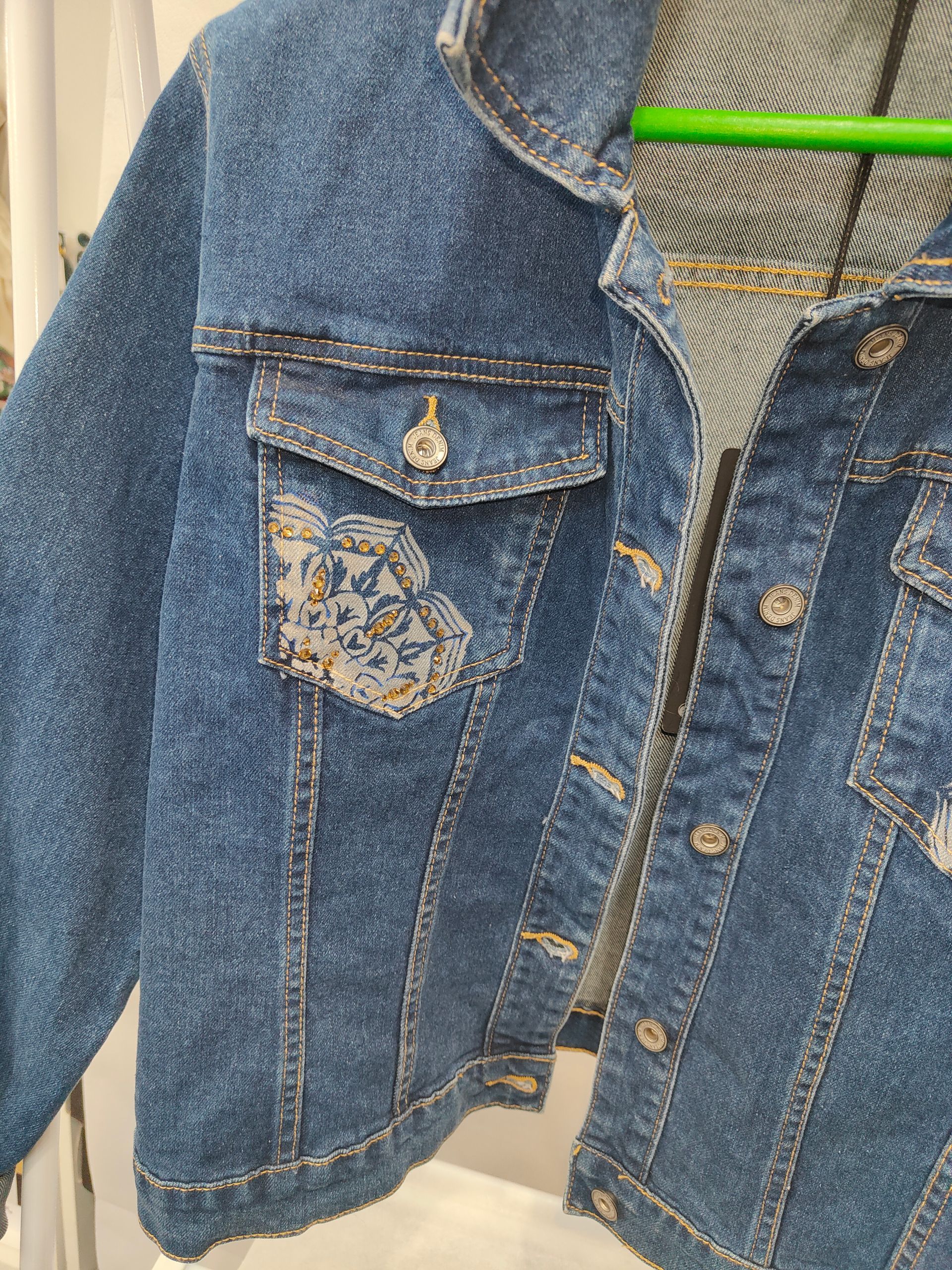 Una giacca di jeans blu a lavaggio medio con una toppa in tessuto fantasia sulla tasca sul petto, appesa a una gruccia verde.