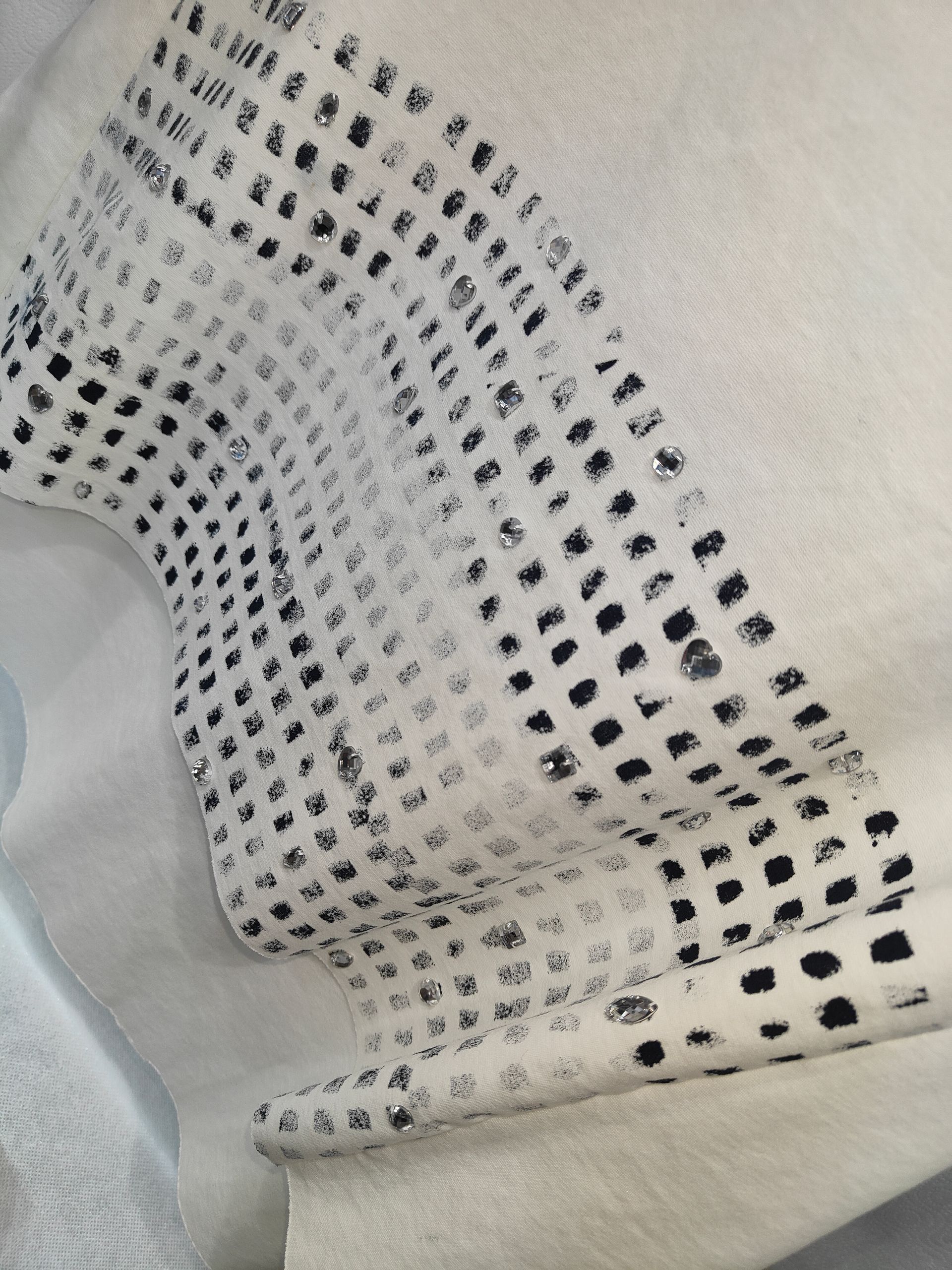 Primo piano di un tessuto color crema con un motivo di piccoli pois neri stampati, impreziositi da strass trasparenti sparsi.