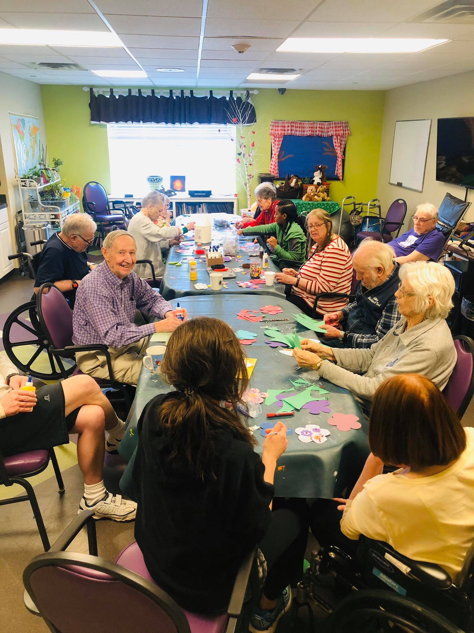 A group of people sits around tables in a brightly lit room working on colorful craft projects.
