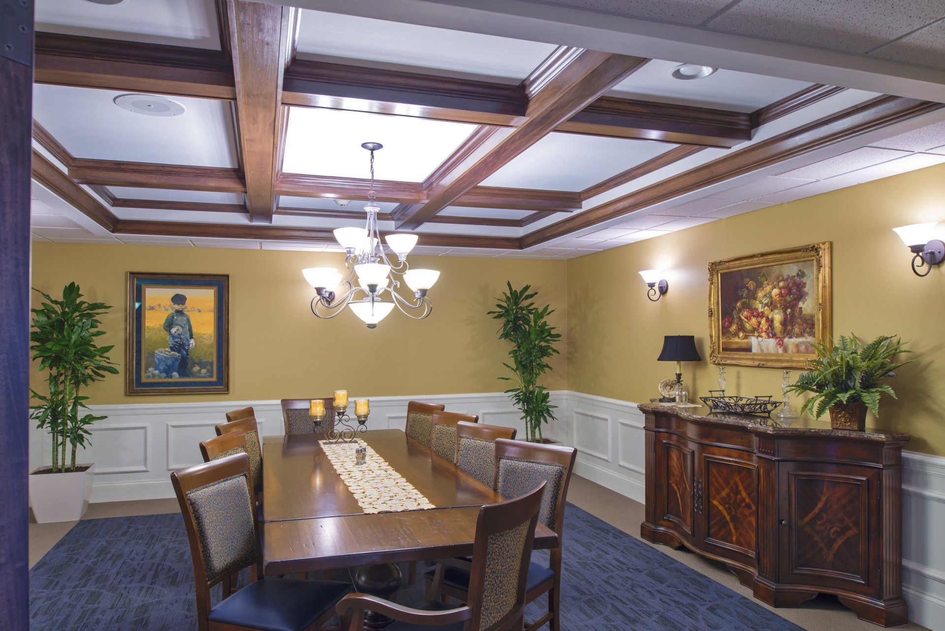 A formal dining room features a long wooden table, chandelier, coffered ceiling, yellow walls, and decorative artwork.