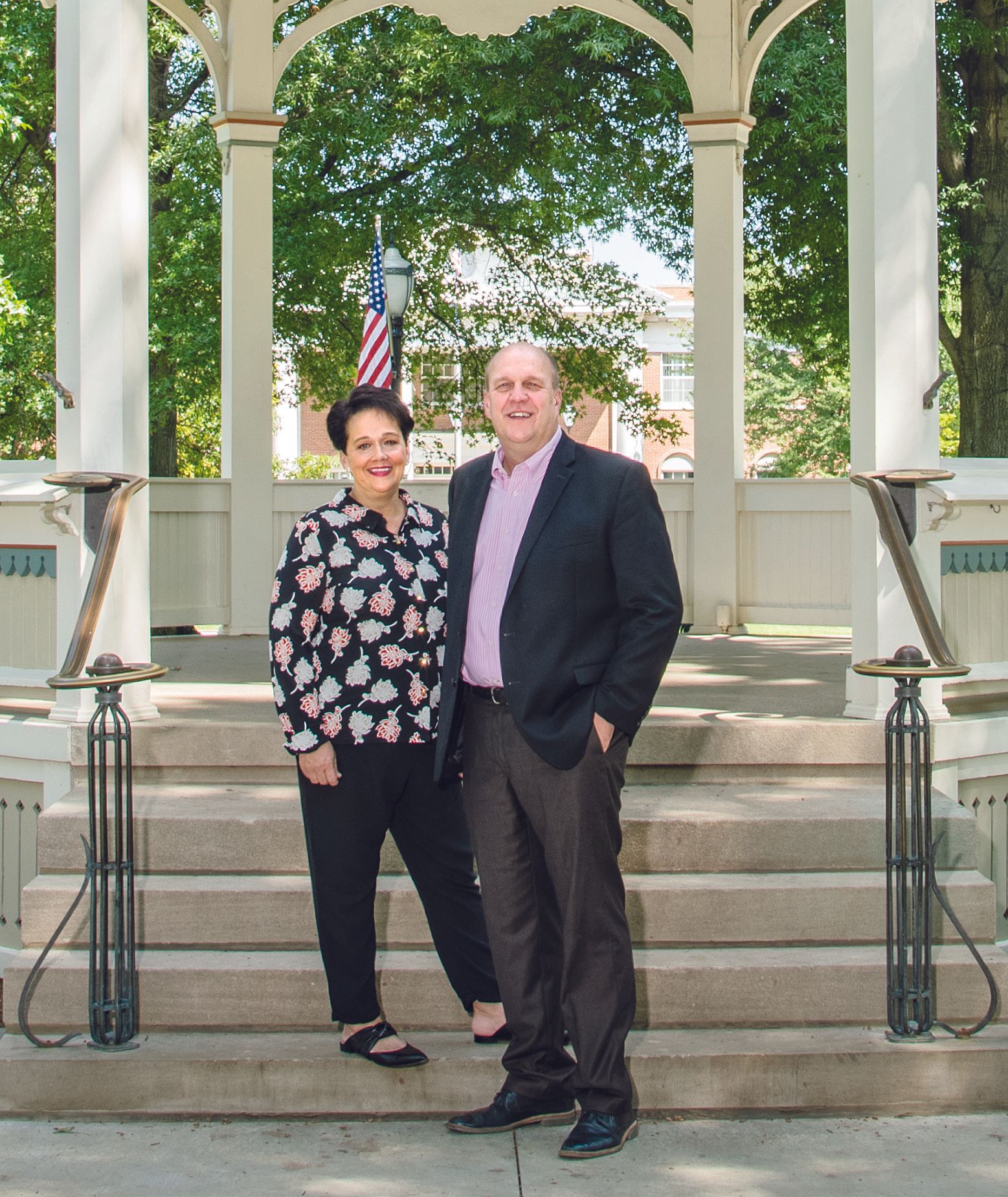 Two people stand smiling on the stairs of a white gazebo with a flag in the background.