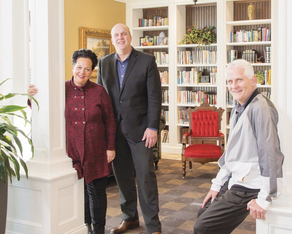 Three people stand and pose in a library featuring bookshelves and a red chair.