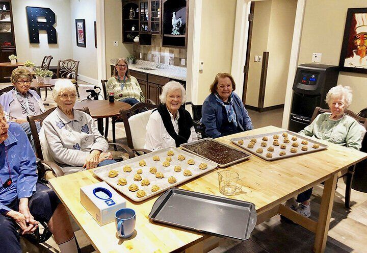 Six people sit at a table covered in trays of cookies and baking sheets in a communal activity space.